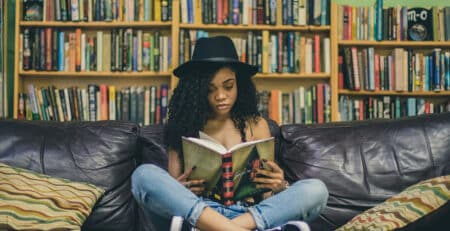 A woman reads a book on a couch next to a bookshelf