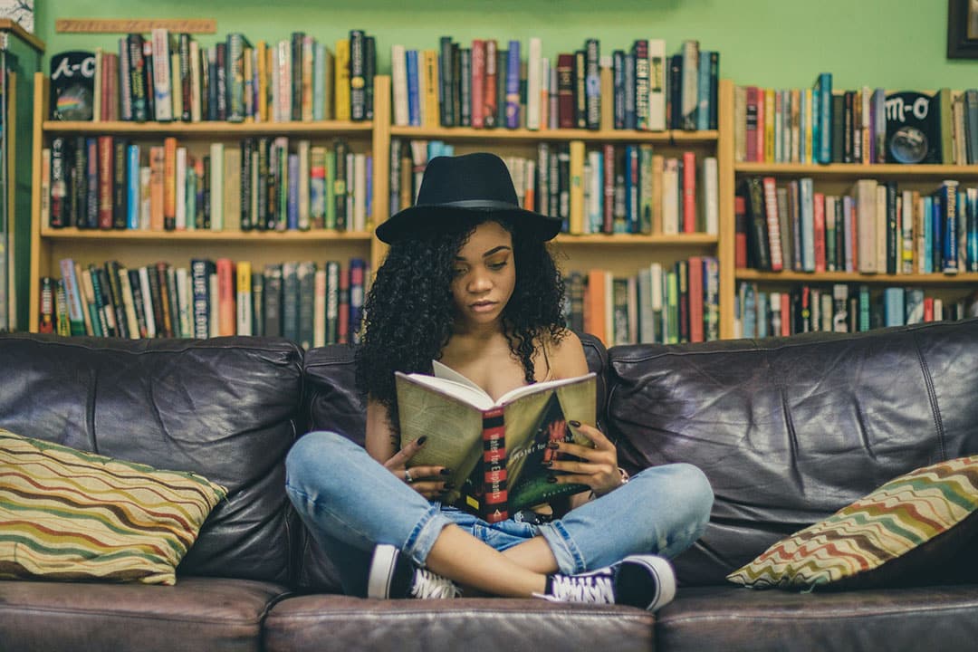 A woman reads a book on a couch next to a bookshelf